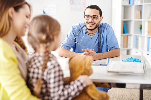 Portrait of friendly Middle-Eastern pediatrician talking to little girl and his mom smiling happily during consultation in doctors office