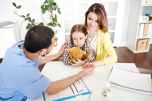 Back view portrait of young doctor talking to scared little girl hugging teddy bear trying to help her calm down during consultation in modern clinic