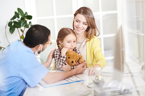 Back view portrait of young doctor talking to scared little girl hugging teddy bear sitting in mothers lap during consultation in modern clinic