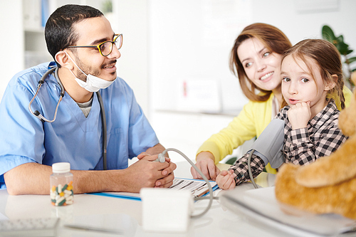 Portrait of cheerful Middle-Eastern pediatrician measuring blood pressure of cute little girl sitting in her mothers lap during consultation in modern clinic