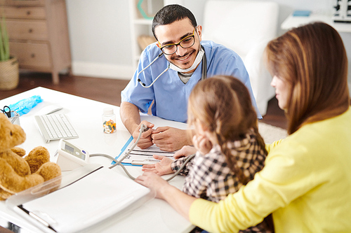 Portrait of cheerful Middle-Eastern pediatrician smiling to cute little girl sitting in her mothers lap during consultation in modern clinic