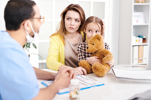 Portrait of worried young mother and cute little girl visiting doctor and talking to pediatrician during consultation