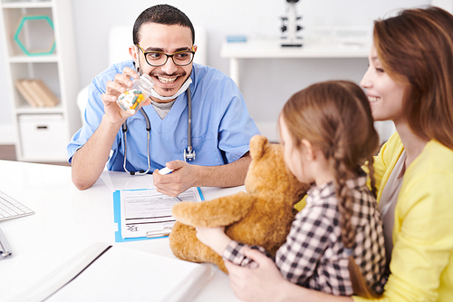 Portrait of playful Middle-Eastern pediatrician offering vitamins to little girl and smiling happily during consultation in modern clinic