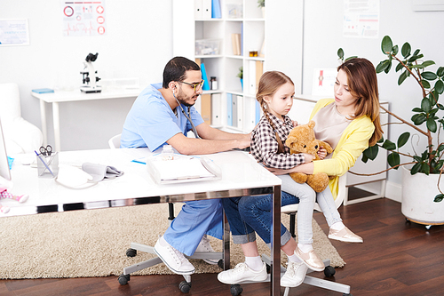 full length portrait of young middle-. doctor listening to child breathing from the back using stethoscope during consultation in modern clinic