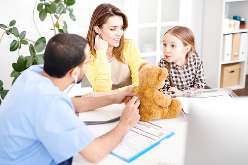 back view portrait of young middle-. doctor putting stethoscope to plush teddy bear toy during consultation with little girl and her mother in modern clinic.