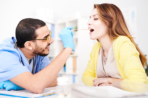 side view portrait of young middle-. doctor examining throat of beautiful young woman, asking her to open mouth during consultation in modern clinic