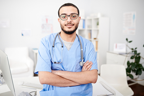 Waist up portrait of confident Middle-Eastern doctor wearing glasses posing in modern office standing with arms crossed and 