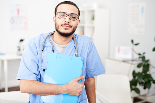 Waist up portrait of young Middle-Eastern doctor wearing glasses posing in modern office holding clipboard  and , copy space