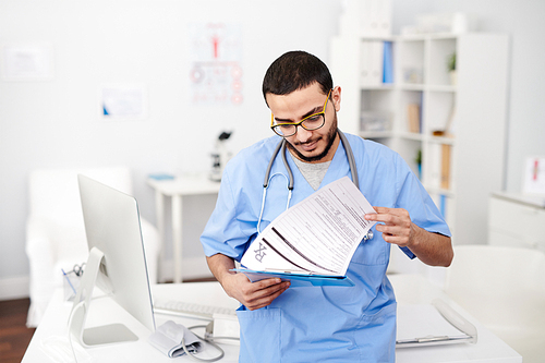 Waist up portrait of young Middle-Eastern doctor wearing glasses posing standing by desk in modern office and reading patients report, copy space