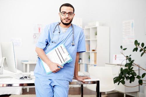 Portrait of young Middle-Eastern doctor wearing glasses posing standing by desk in modern office holding clipboard  and , copy space