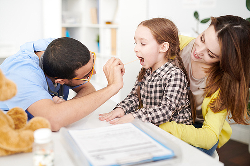 Side view portrait of cute little girl visiting doctor and sitting in her mothers lap with mouth open during examination