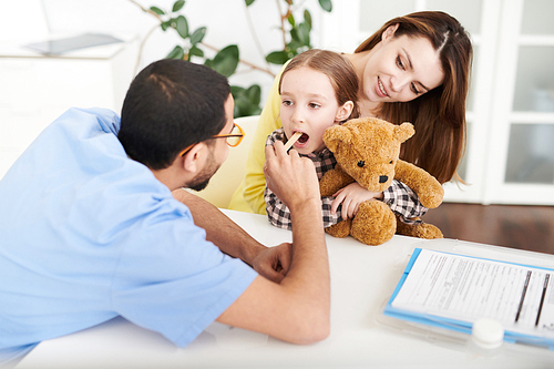 Portrait of cute little girl visiting doctor and sitting in her mothers lap with mouth open during examination