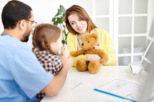 Portrait of beautiful young mother playing with cute little girl trying to calm her down  during visit to doctors office in modern clinic