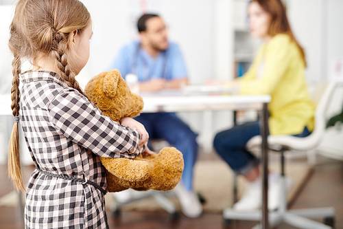 Back view portrait of scared little girl standing in doorway of doctors office and hugging teddy bear toy, looking at mom and pediatrician, copy space