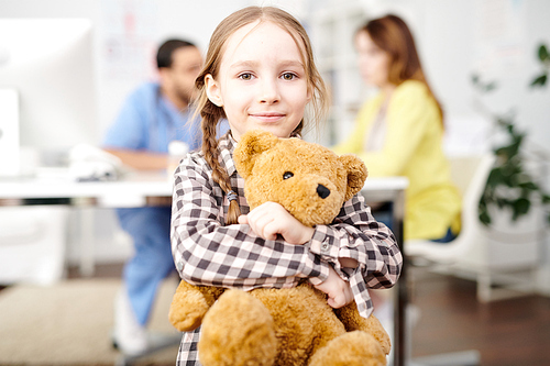 Portrait of cute little girl posing in doctors office smiling happily and hugging teddy bear while 