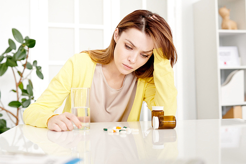 Portrait of frustrated  young woman sitting at desk in doctors office and looking at many assorted pills