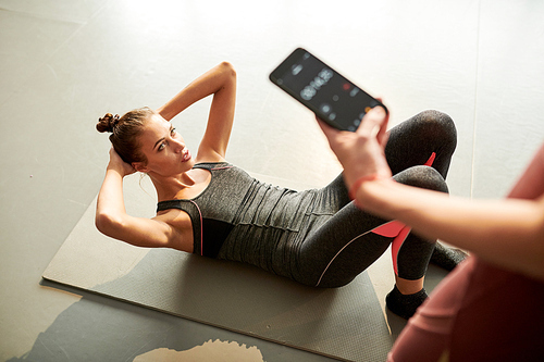 Above view portrait of contemporary young woman doing crunches during fitness workout in health club while female instructor holding smartphone with timer