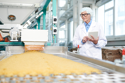 Waist up portrait of senior woman working at food factory standing by conveyor belt and using digital tablet during quality inspection, copy space