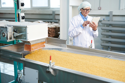 Waist up portrait of smiling senior woman working at factory standing by conveyor belt and using digital tablet at food production, copy space