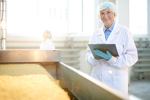 Portrait of smiling  senior woman working at factory and controlling food production standing by macaroni conveyor belt, copy space