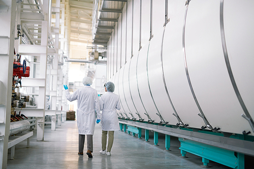 Back view full length portrait of two factory workers wearing white coats crossing production workshop, copy space