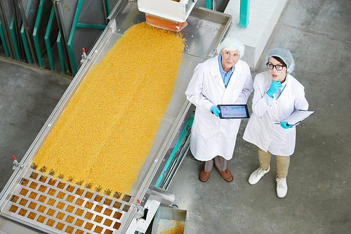 High angle  portrait of two female factory workers standing by macaroni conveyor belt and looking up during quality inspection at food production, copy space