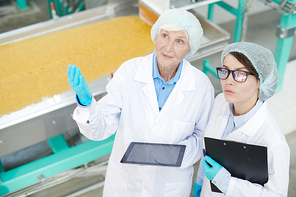 High angle  portrait of two female factory workers standing by macaroni conveyor belt and looking up during quality inspection at food production, copy space