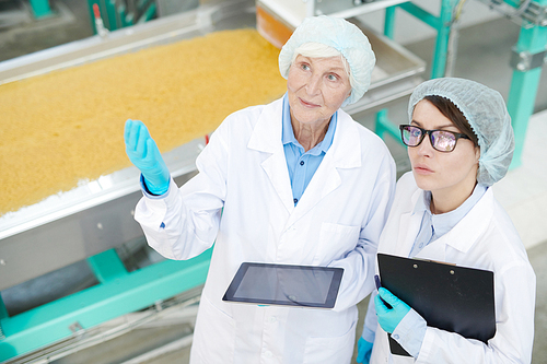 High angle  portrait of two female factory workers standing by macaroni conveyor belt and looking up during quality inspection at food production, copy space