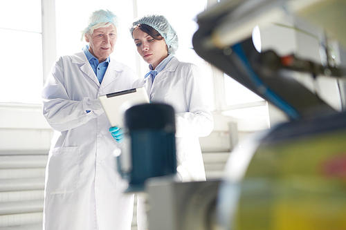 Portrait of senior factory worker wearing clean white coat standing by machine units at packaging line and talking to young trainee, copy space