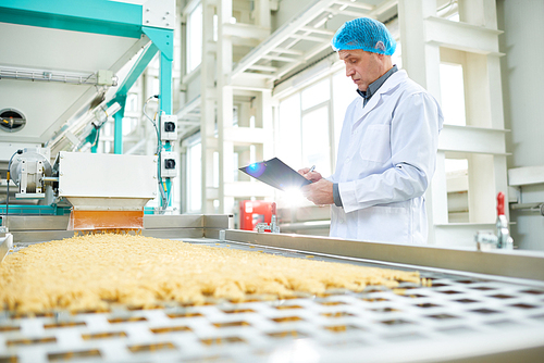 Side view portrait of senior factory worker  in food industry holding clipboard standing by conveyor belt, copy space