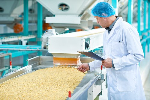 Side view portrait of senior factory worker doing  production quality inspection in food industry holding clipboard standing by conveyor belt, copy space