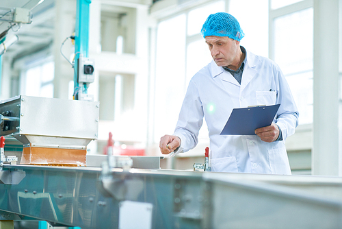 Waist up portrait  of  senior factory worker doing  production quality inspection holding clipboard and making notes standing by conveyor belt in modern production workshop, copy space