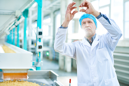 Waist up portrait of senior factory worker doing quality inspection in food industry holding two macaronis standing by conveyor belt, copy space