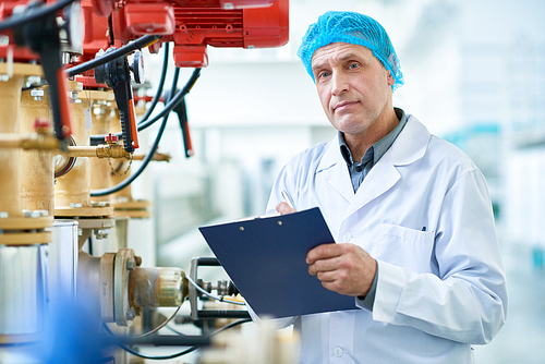 Waist up portrait  of senior factory worker doing  production quality inspection standing by  pipes and valves in modern workshop holding clipboard, copy space