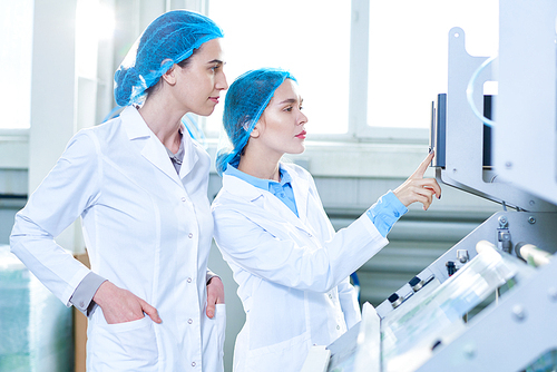 Side view portrait of two female specialists wearing lab coats working at modern food factory and checking production process standing by power units, copy space