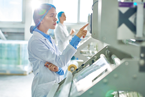 Side view portrait of young female worker wearing lab coat and hair cover standing by  power units pressing buttons on control panel in clean production workshop.