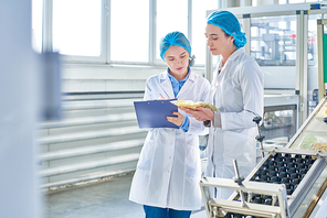 Portrait of  two young female workers wearing lab coats and hair covers standing by  power units and writing on clipboard  in clean production workshop, copy space