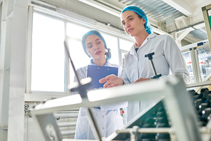 Low angle portrait of  two young female workers wearing lab coats standing by  power units and writing on clipboard  in clean production workshop, copy space