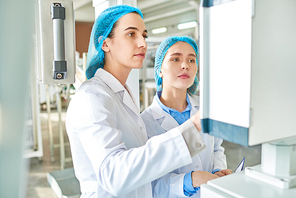 Waist up  portrait of  two young female workers wearing lab coats standing by  power units and pressing buttons on control panel  in clean production workshop, copy space