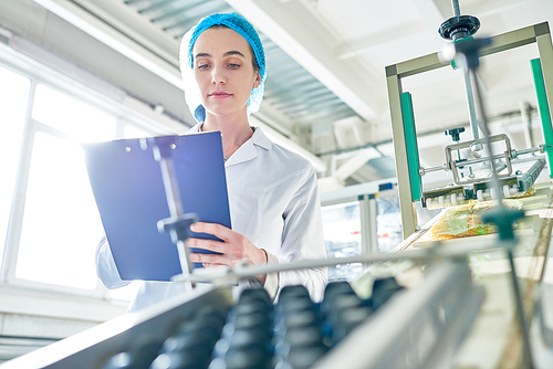 Low angle portrait of  young female worker wearing lab coat standing by  power units and writing on clipboard  in clean production workshop, copy space