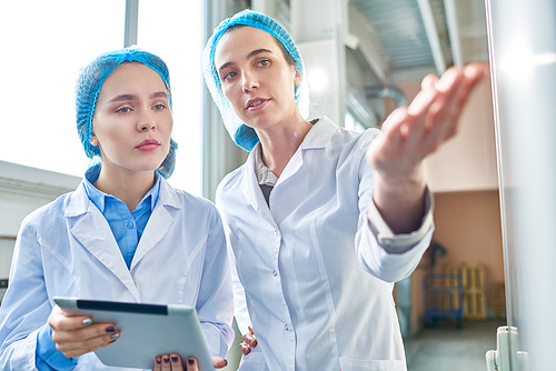 Waist up portrait of  two young female workers wearing lab coats standing by  power units and discussing work standing  in clean production workshop, copy space