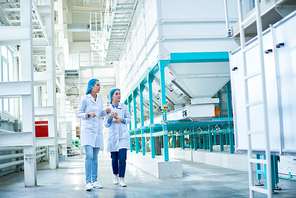 Full length portrait of  two young female workers wearing lab coats walking by  power units crossing hall in  in clean production workshop, copy space