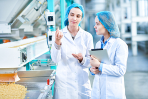 Portrait of  two young female workers wearing lab coats standing by  conveyor line with macaroni  in clean production workshop
