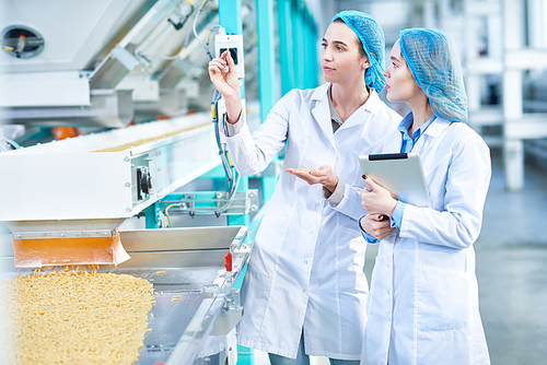 Portrait of  two young female workers wearing lab coats standing by  conveyor line with macaroni inspecting quality in clean production workshop, copy space