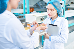 Waist up portrait of  two young female workers wearing lab coats standing by  conveyor line with macaroni inspecting quality in clean production workshop, copy space