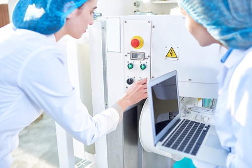 Back view portrait of  two young female workers wearing lab coats setting up power units and pushing buttons on control panel and using laptop in clean production workshop, copy space