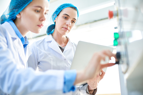 Low angle portrait of  two young female workers wearing lab coats setting up power units and pushing buttons on control panel in clean production workshop, copy space