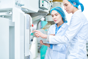 Side view portrait of  two young female workers wearing lab coats setting up power units and pushing buttons on control panel in clean production workshop, copy space