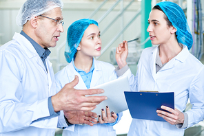 Waist up portrait of two young women asking questions to senior factory worker during training seminar for new specialists standing in clean workshop of modern plant.