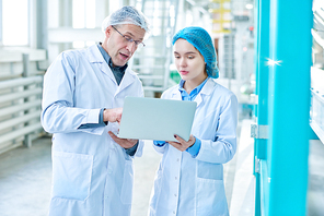 Portrait of senior factory worker wearing lab coat training young woman and using laptop while standing in clean production workshop of modern plant